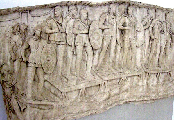 Fanteria ausiliaria romana che attraversa un fiume, probabilmente il Danubio, su un ponte di barche durante le guerre dei Daci dell'imperatore Traiano (101–106 d.C.). Si distinguono per lo scudo ovale (clipeus) di cui erano dotati, in contrasto con lo scutum rettangolare portato dai legionari. Pannello della Colonna Traiana, Roma